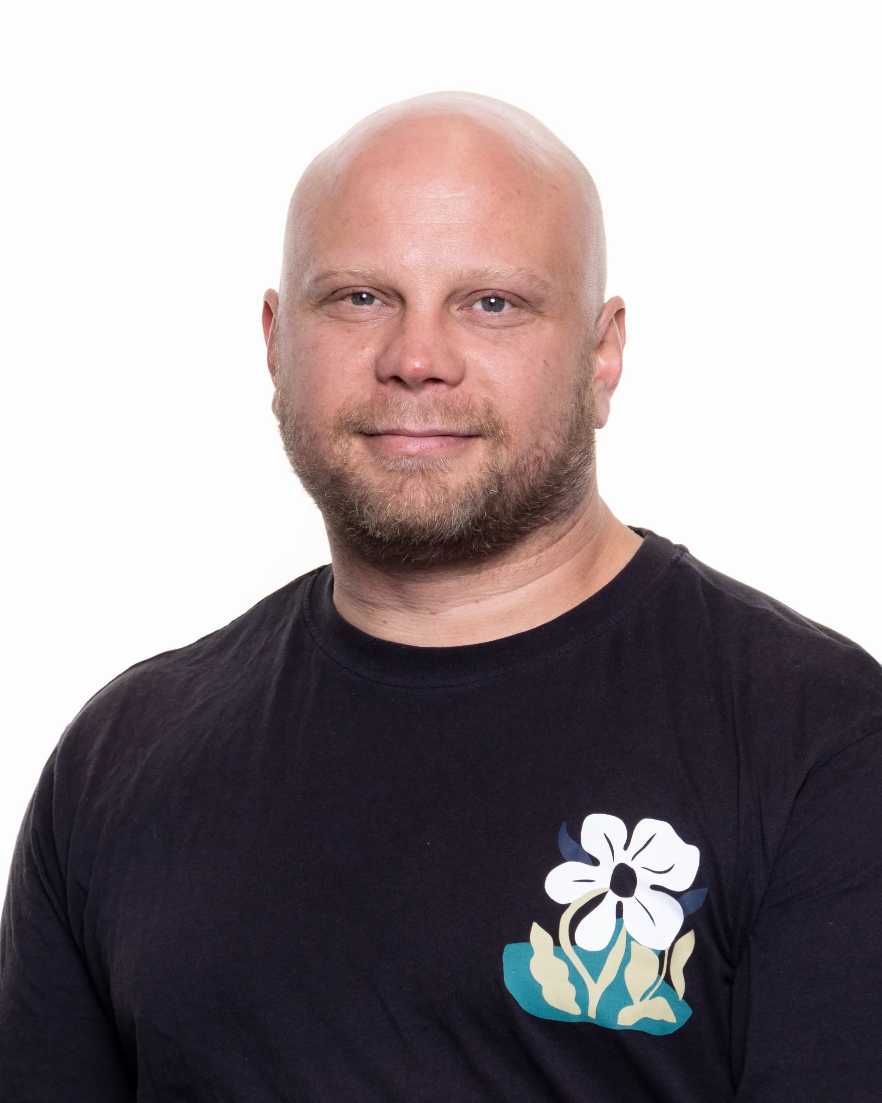 Valkopaitainen kalju mies, jolla on parta ja kukka-aiheinen kuvio paidassaan, valkoisella taustalla.