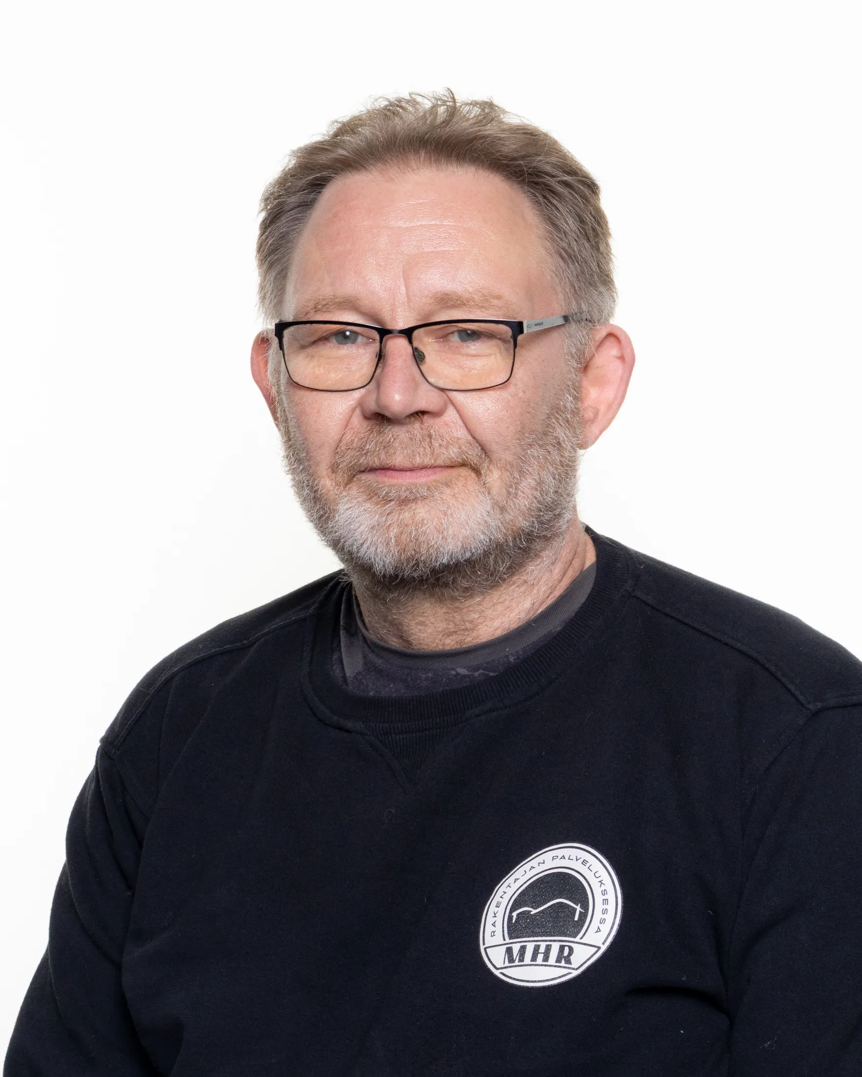 Lähikuva keski-ikäisestä miehestä, jolla on harmaat hiukset ja parta, silmälasit ja musta pusero, jossa on valkoinen logo. Kuva on otettu valkoista taustaa vasten.