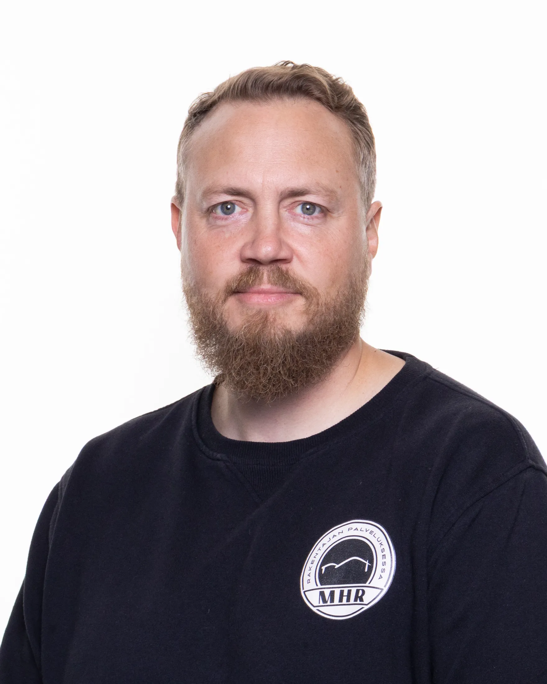 Muotokuva miehestä, jolla on parta, siniset silmät ja musta pusero, jossa on MHR-logo valkoisella taustalla. Kuva on otettu valkoisella taustalla, ja valaistus on tasainen.