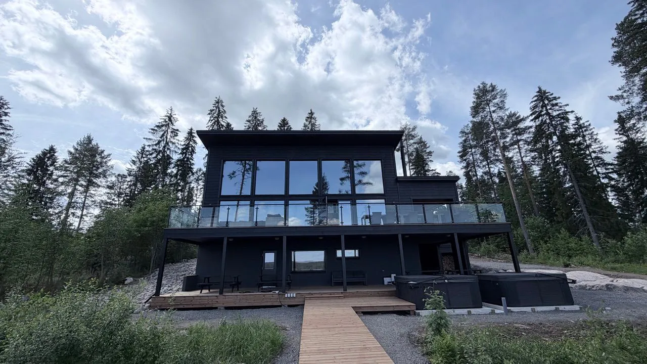 Moderni, tumma kaksikerroksinen talo metsäisessä maisemassa. Talossa on suuret ikkunat, jotka heijastavat pilvistä taivasta ja havupuita. Yläkerrassa on lasikaiteellinen parveke ja alakerrassa puinen terassi, jolle johtaa puinen kävelysilta. Terassilla on ulkokalusteita ja kaksi poreallasta. Kuva on otettu kirkkaassa, puolipilvisessä säässä.
